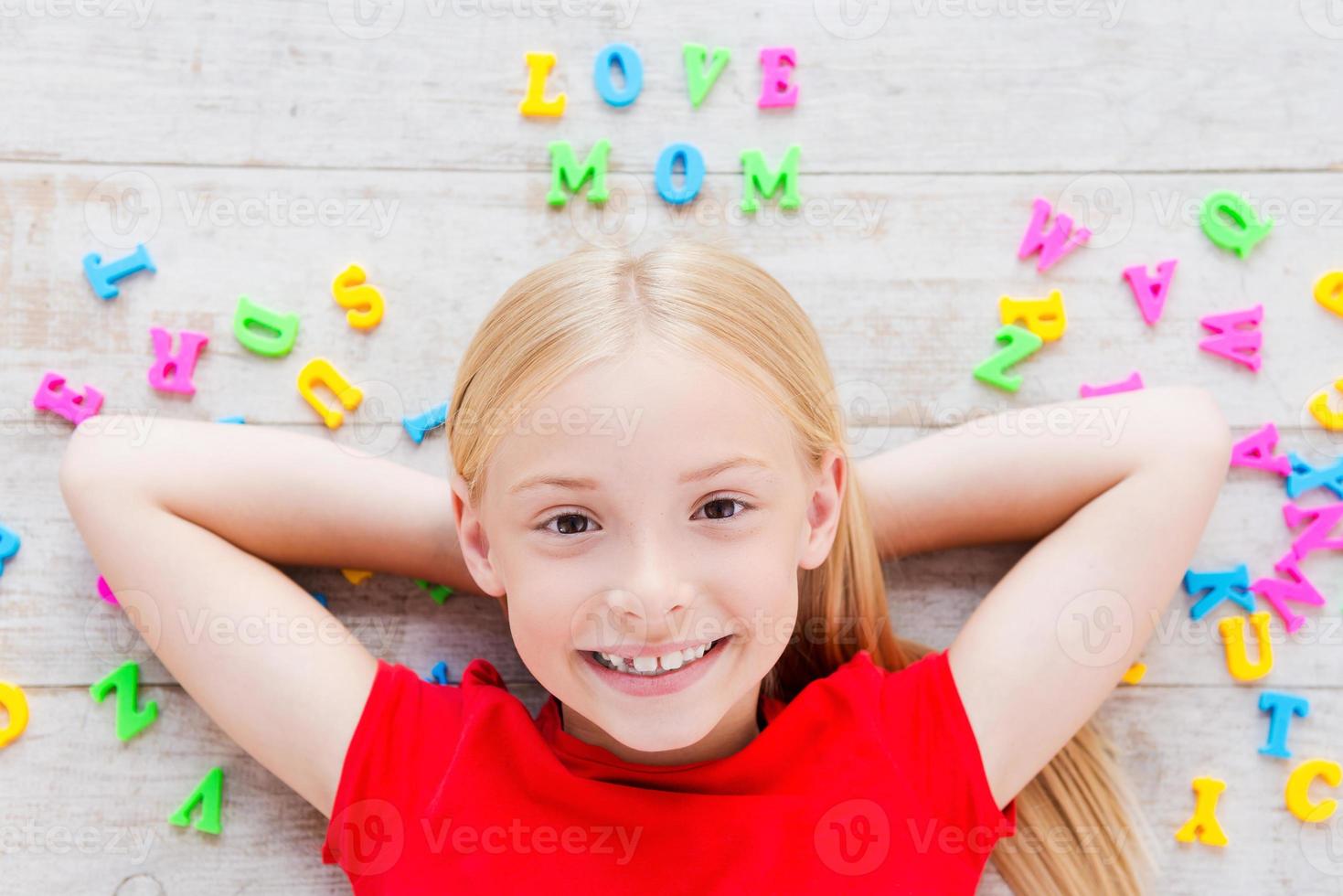 I love my mom Top view of cute little girl holding hands behind head while lying on the floor with plastic colorful letters laying around her photo