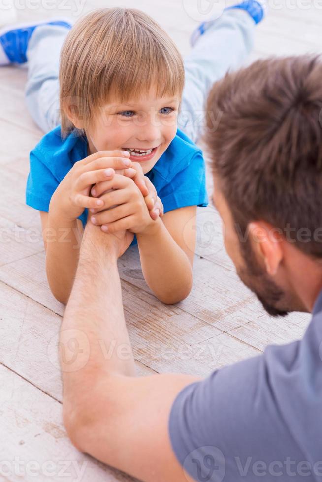 I want to be as strong as my father Top view of cheerful father and son competing in arm wrestling while both lying on the hardwood floor photo