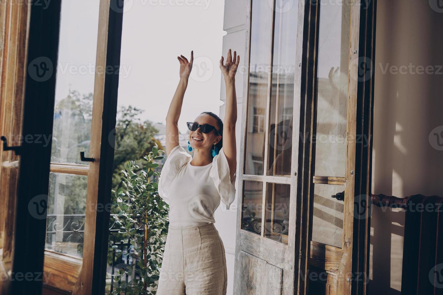 Fashionable young woman keeping arms raised and smiling while standing on the balcony photo