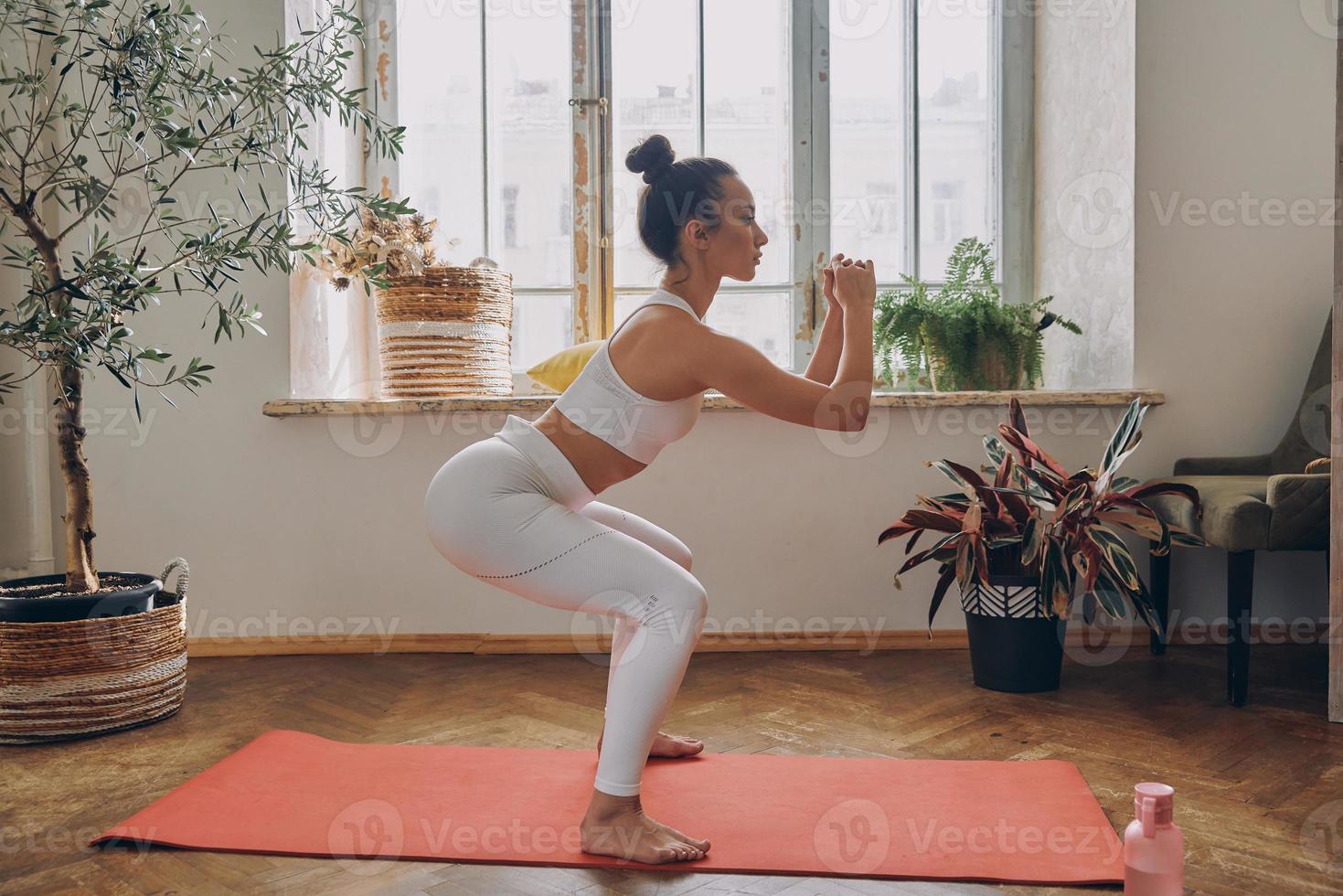 Confident woman standing in squatting position while practicing in front of the window at home photo
