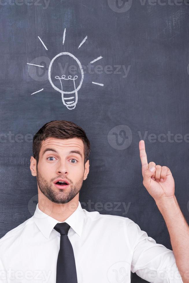 I have an idea Handsome young man pointing up while standing against blackboard with chalk drawing of light bulb photo
