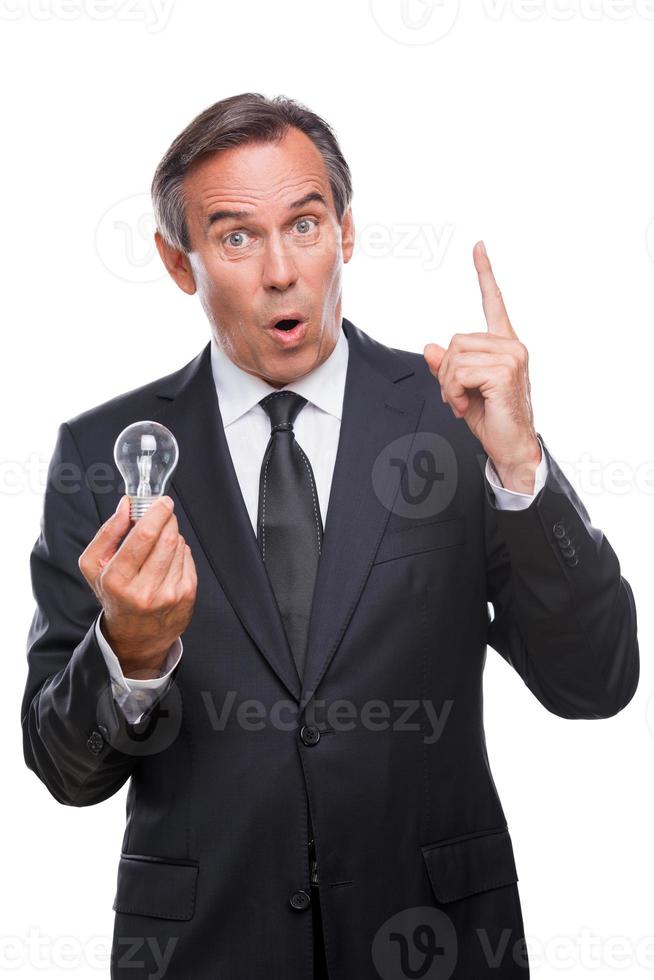 He got an idea  Excited mature man in formalwear holding light bulb and pointing up while standing isolated on white background photo