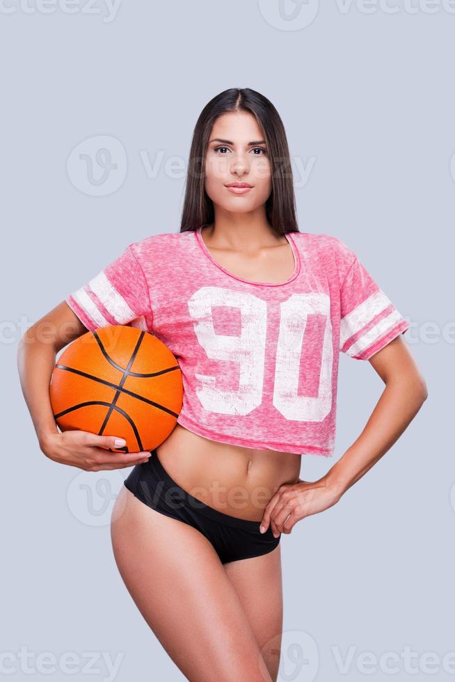 I love basketball Attractive young female cheerleader holding basketball ball and looking at camera while standing against grey background photo