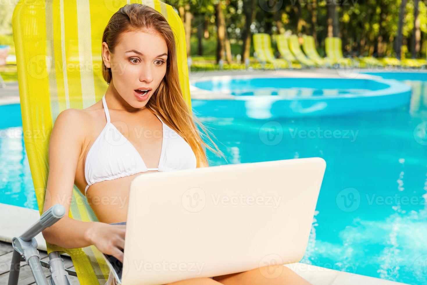 es simplemente increíble y hermosa mujer joven en bikini blanco mirando la computadora portátil y manteniendo la boca abierta mientras se sienta en la tumbona junto a la piscina foto