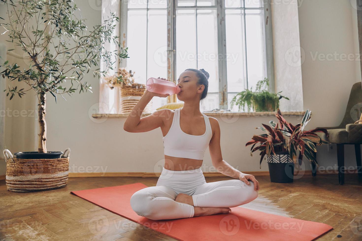 Beautiful young woman drinking water after training while sitting on the exercise mat at home photo