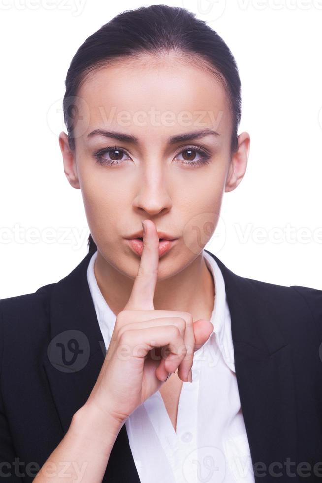 Keep silence Confident young woman in formalwear looking at camera and holding finger on lips while standing isolated on white background photo
