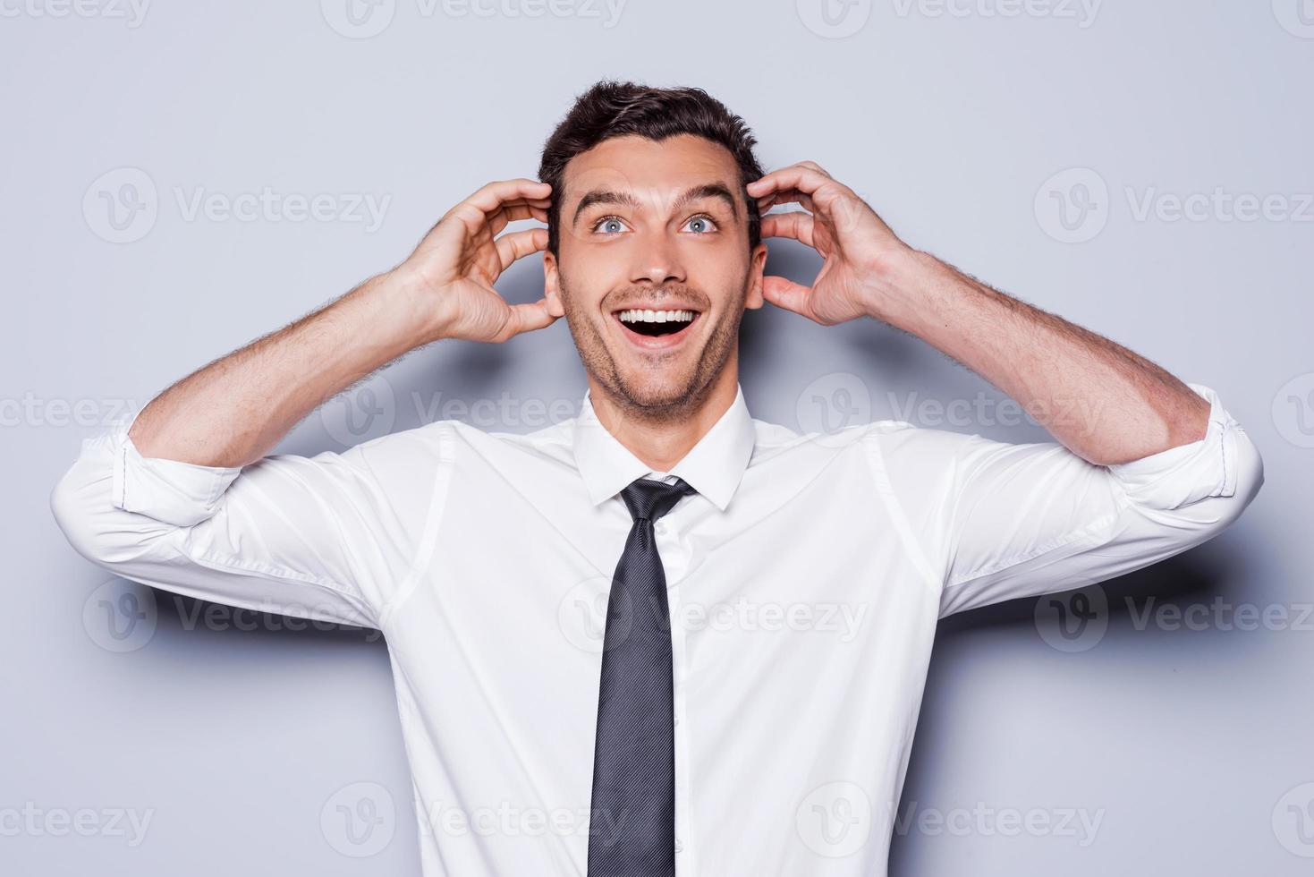 It is just unbelievable Happy young man in shirt and tie touching head with hands and smiling while standing against grey background photo