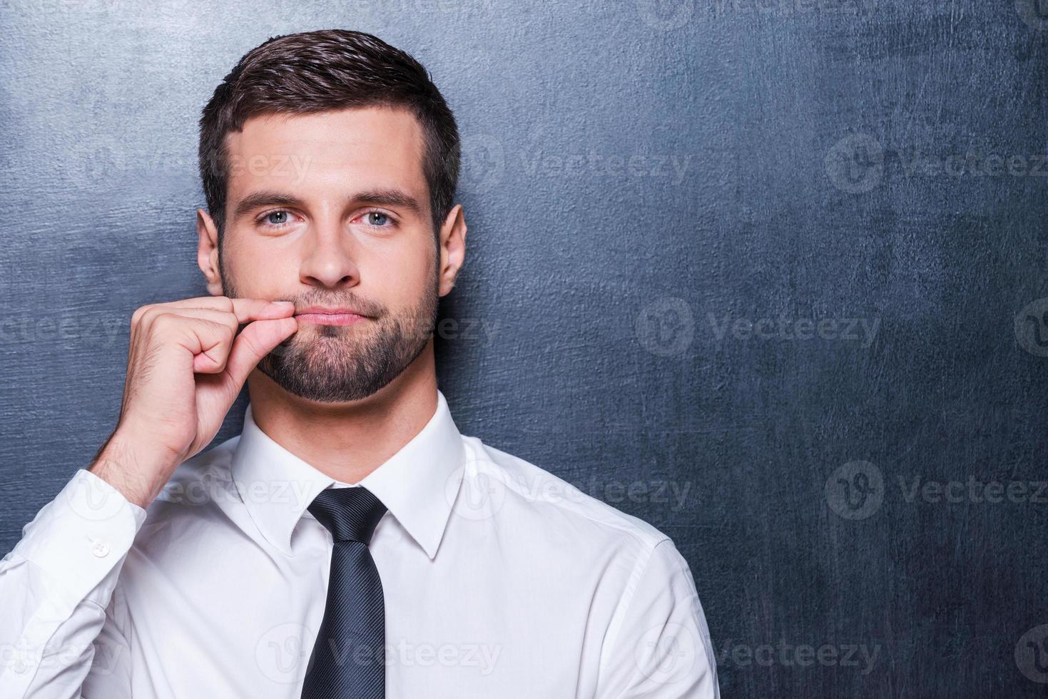Keep silence Handsome young man in formalwear showing a sign of closing mouth and looking at camera while standing against blackboard photo
