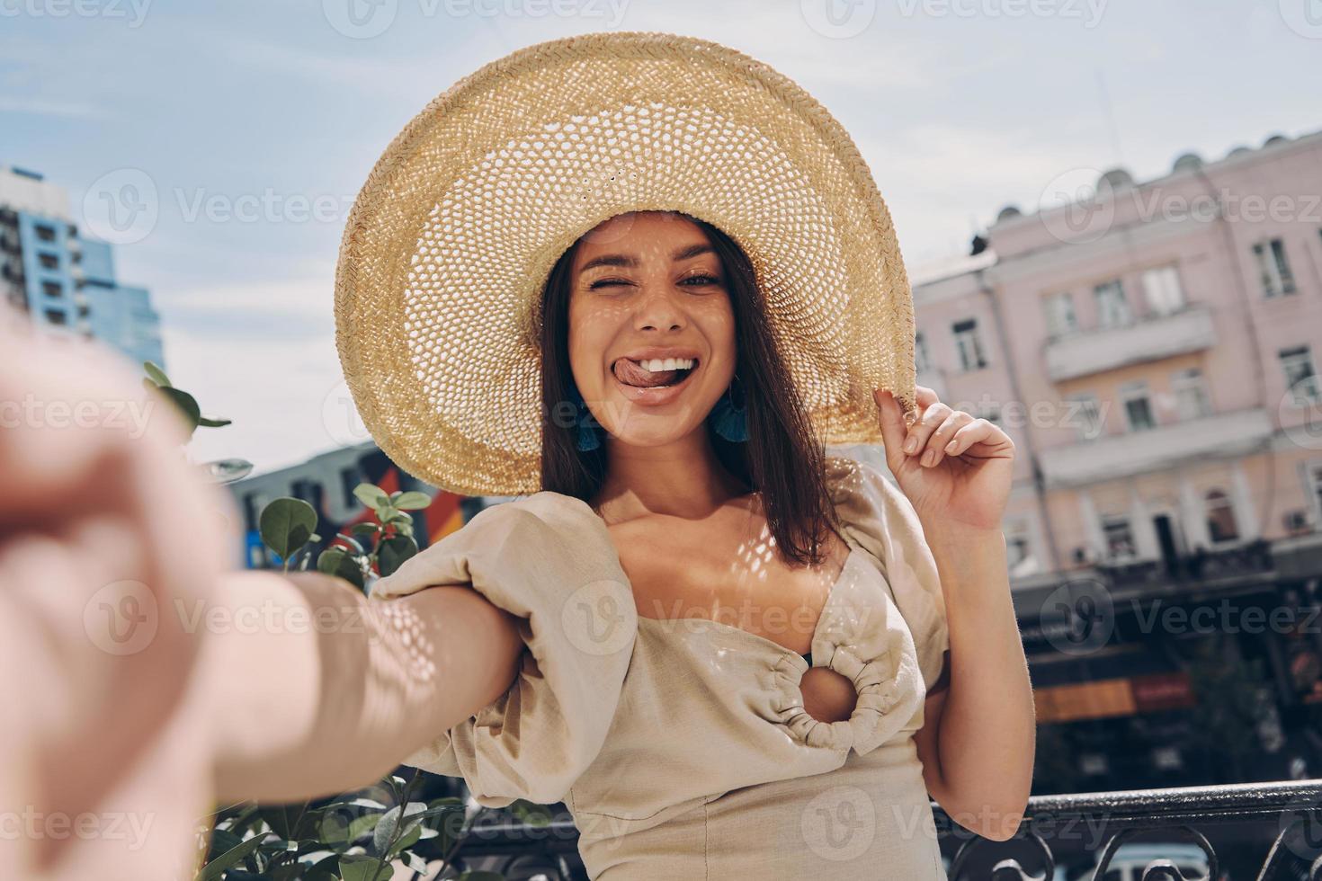 Playful young woman in elegant hat making selfie while relaxing on the balcony photo