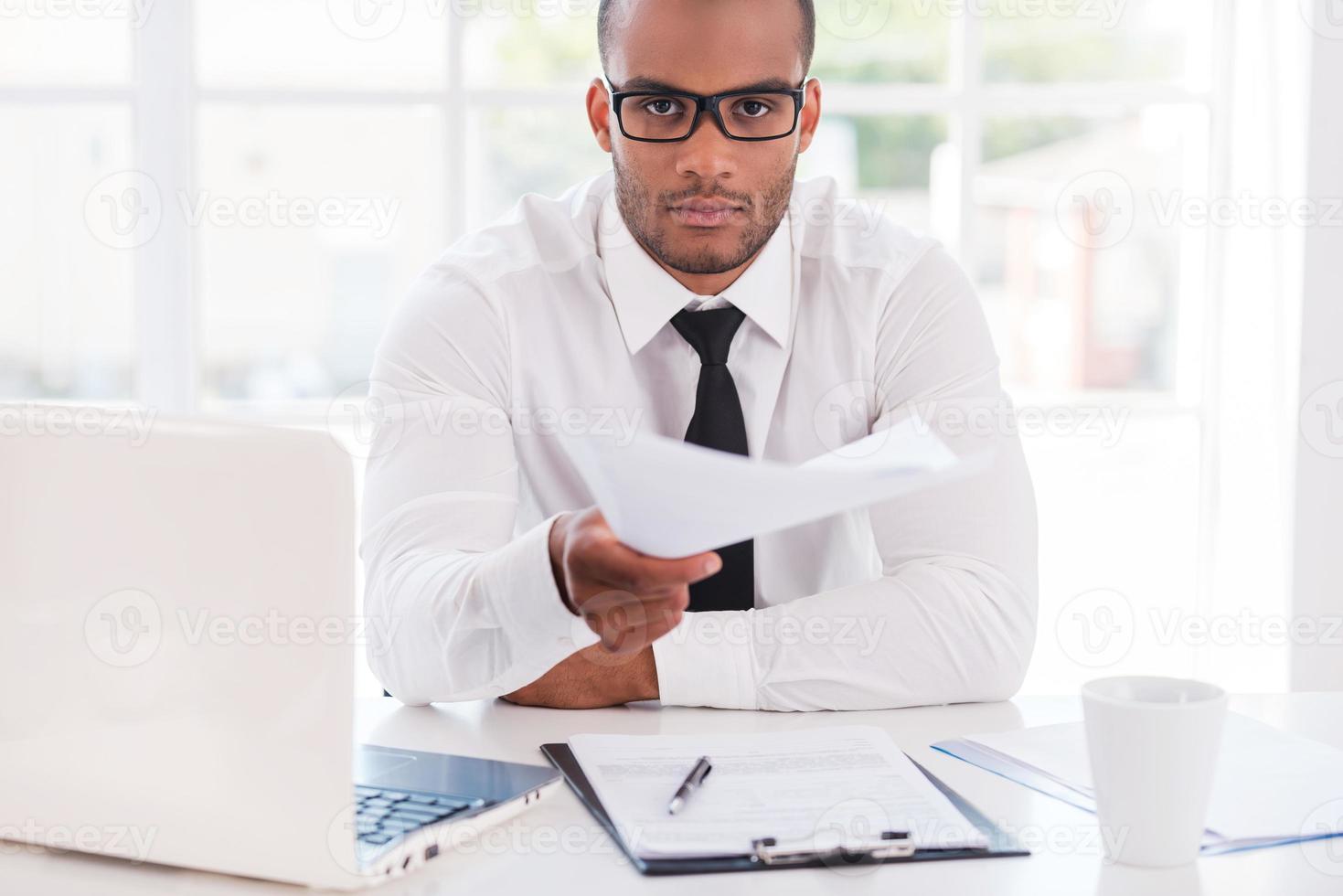 I have some paperwork for you Confident young African man in shirt and tie stretching out documents and looking at you while sitting at his working place photo