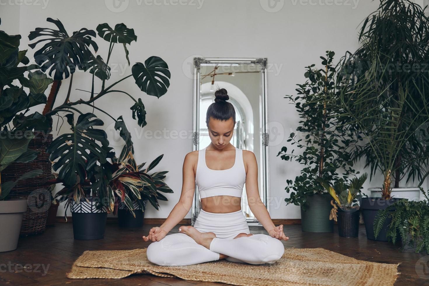 mujer joven concentrada meditando mientras se sienta en el suelo con plantas de interior a su alrededor foto