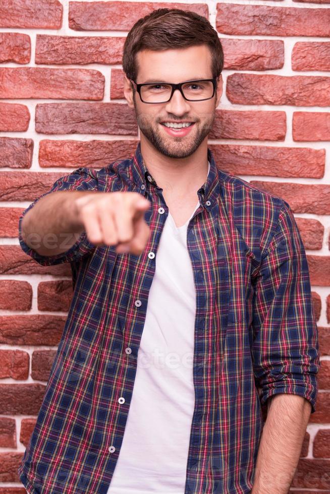 Te elijo a ti, joven apuesto, apuntándote y sonriendo mientras estás de pie contra la pared de ladrillos. foto