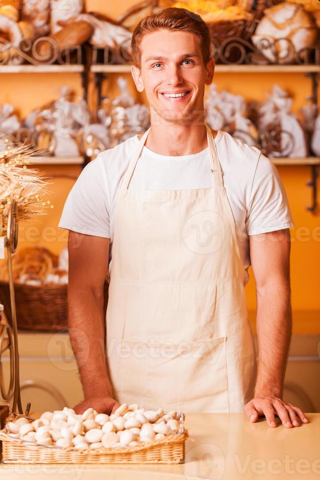 How may I help you Handsome young man in apron looking at camera and smiling while standing in bakery shop photo