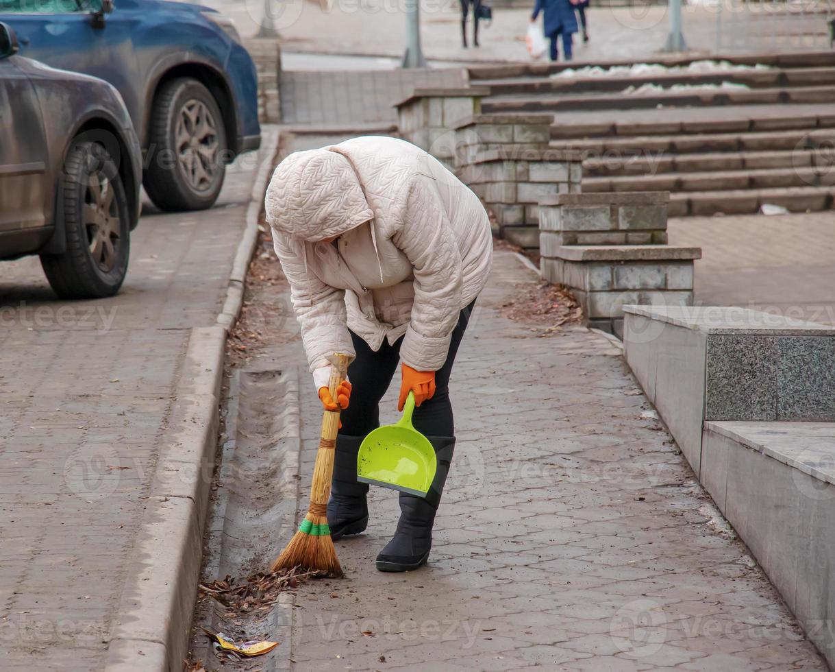 un trabajador con una escoba barre la calle del polvo y los escombros. el conserje limpia el camino. una limpiadora barre la acera con una escoba y un recogedor foto