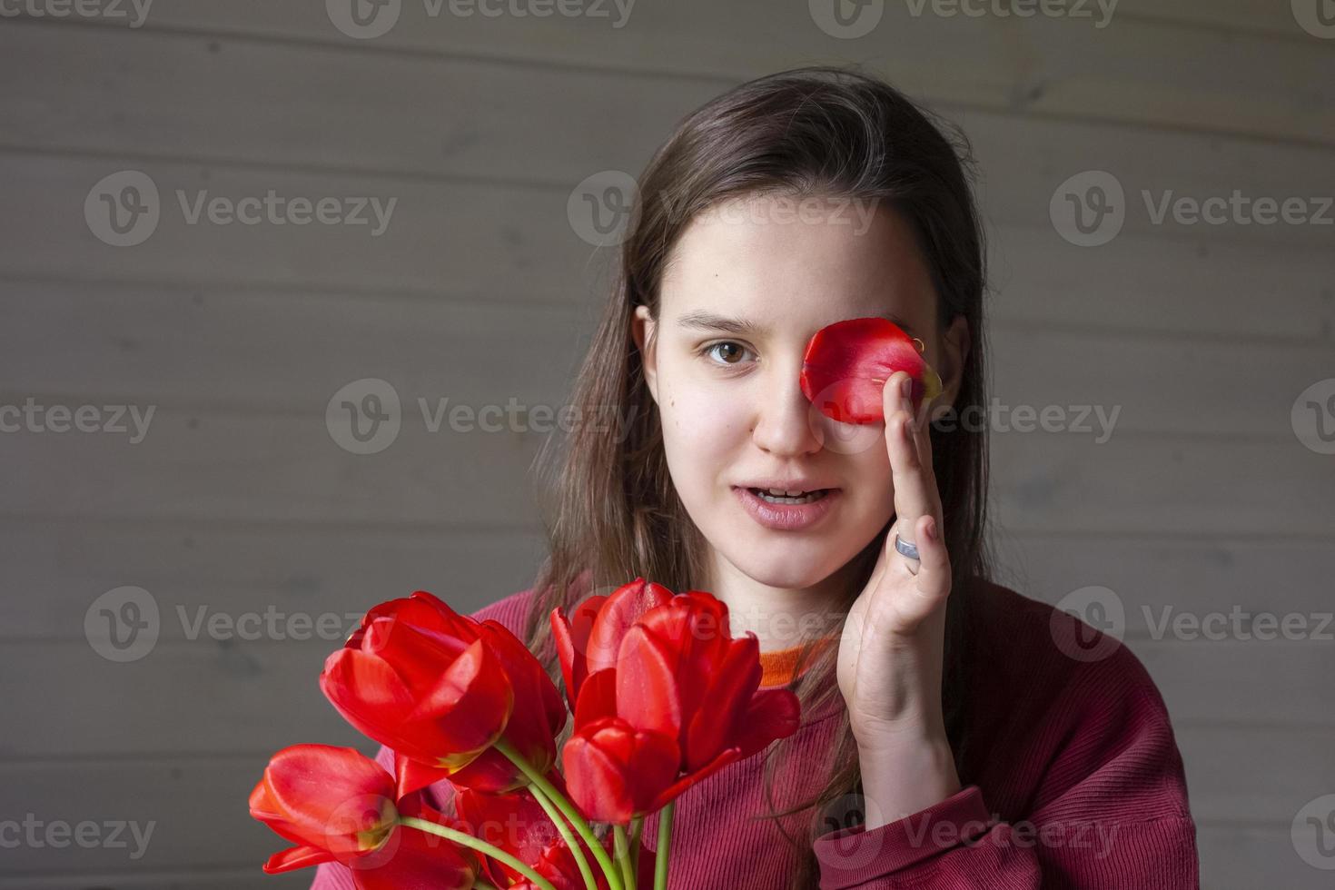 portrait of a brown-haired woman with a bouquet of red tulips on a gray wooden background. a girl in a red pullover with long hair holds a bouquet of tulips in her hands and fools around. Copy space. photo