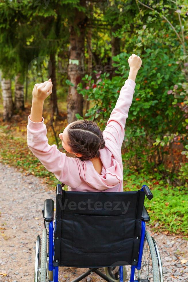 Young happy handicap woman in wheelchair on road in hospital park enjoying freedom. Paralyzed girl in invalid chair for disabled people outdoor in nature. Rehabilitation concept. photo