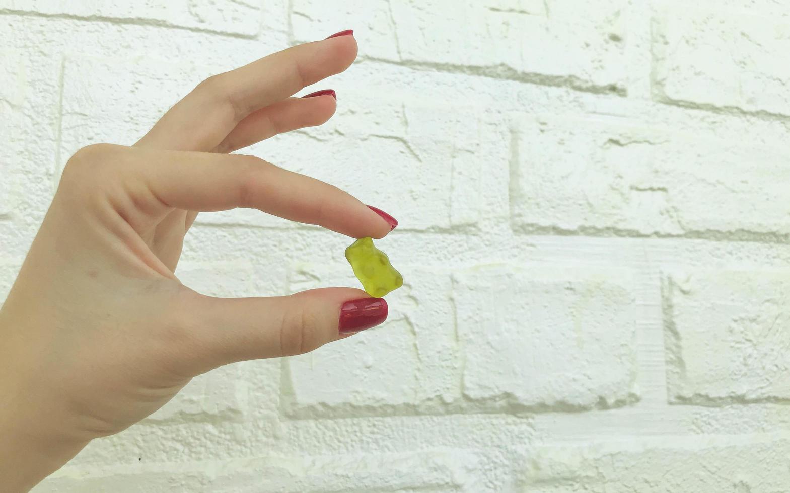 a gummy yellow bear in the hands of a girl with a red manicure. teddy bear is held in hand against the background of a white brick wall made of plaster. sweet candy dessert for tea and coffee photo