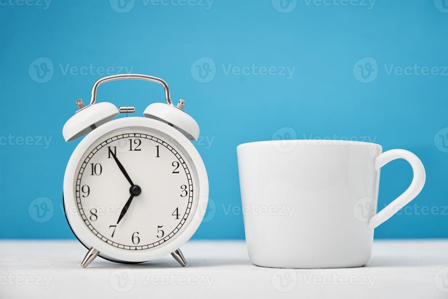 reloj despertador retro blanco y taza sobre fondo azul. concepto de tiempo de la mañana foto