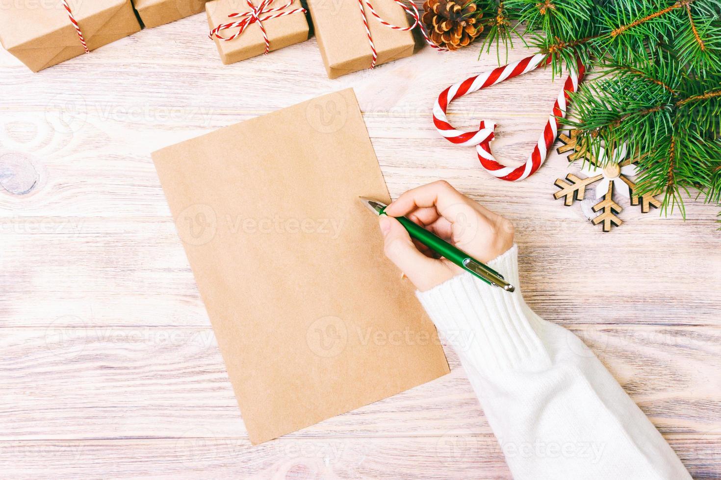 chica mano escribiendo carta de navidad en papel artesanal con decoraciones sobre fondo de madera. tonificado foto