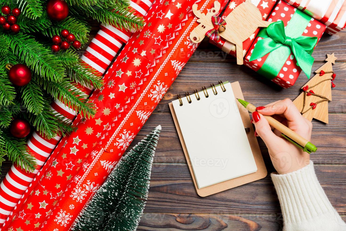 vista superior de la mano femenina escribiendo en un cuaderno sobre fondo de Navidad de madera. abeto y decoraciones festivas. lista de deseos concepto de año nuevo foto