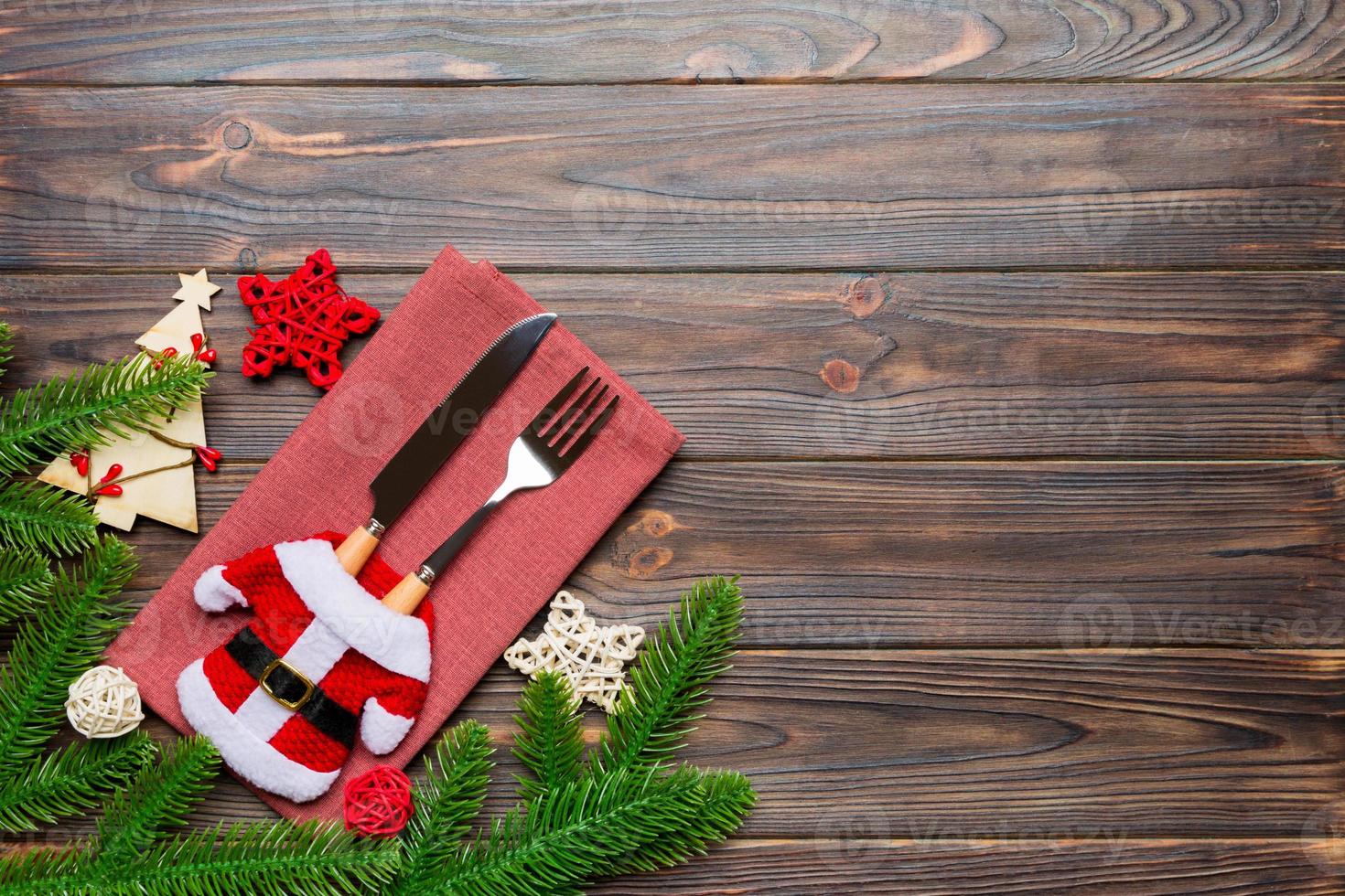 Top view of fork and knife put in santa clothes on napkin with christmas decorations and new year tree on wooden background. Holiday and festive concept with copy space photo