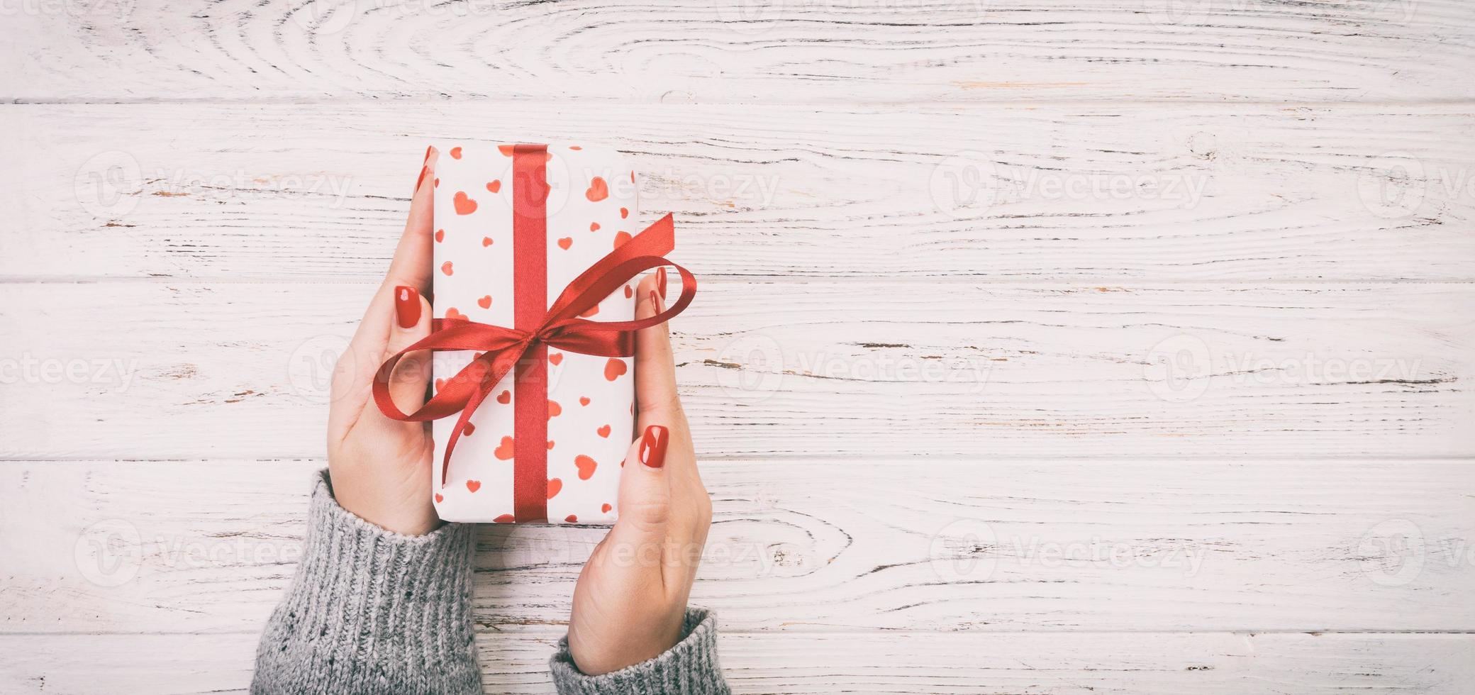 Woman hands give wrapped valentine or other holiday handmade present in paper with red ribbon. Present box, red heart decoration of gift on wooden table, top view with copy space Toned web banner photo