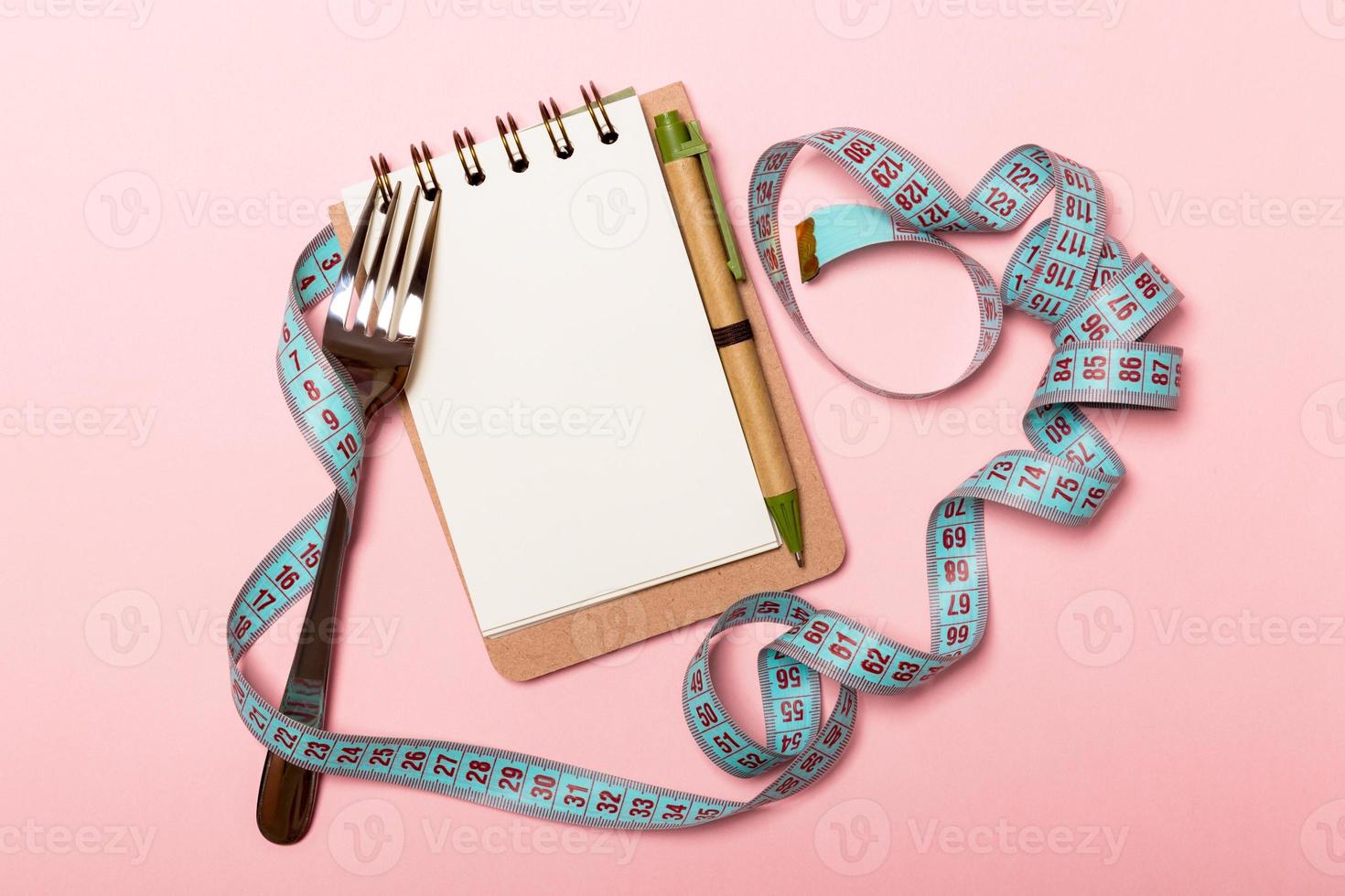 Top view of notebook with fork and blue measuring tape on pink background. Copy space with diet planning photo