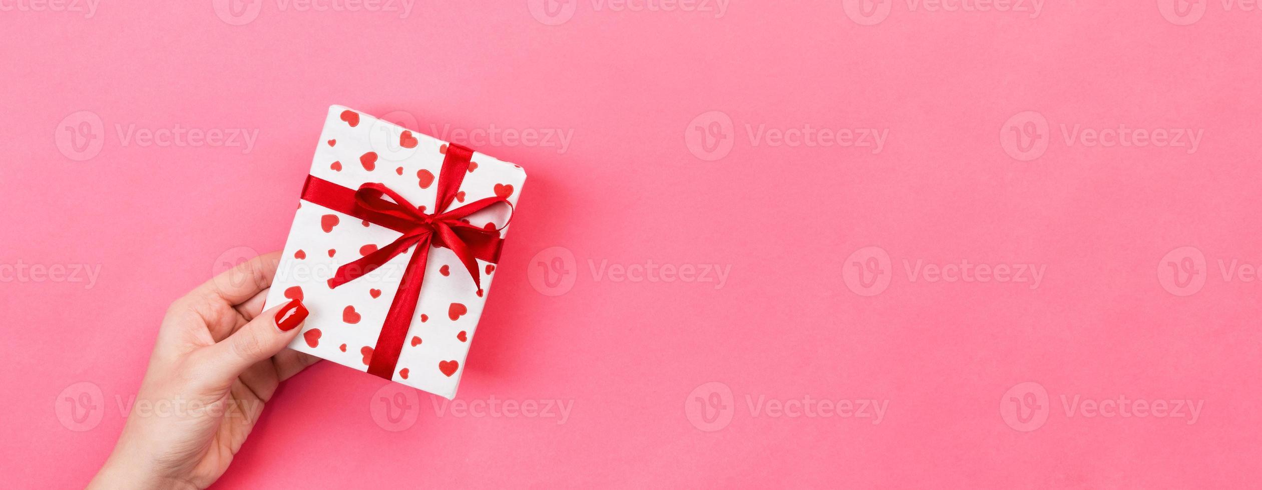Woman hands give wrapped valentine or other holiday handmade present in paper with red ribbon. Present box, red heart decoration of gift on pink table, Top view banner with copy space for you design photo