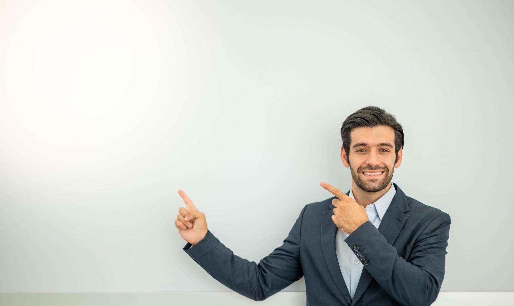 Portrait of smiling young caucasian businessman wearing dark suit and presenting copy space on his pointing finger near with concrete wall. photo