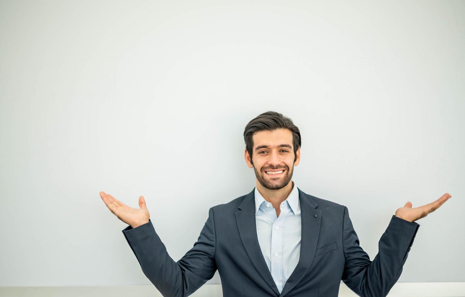 Portrait of smiling young caucasian businessman wearing dark suit and presenting copy space on his palms near with concrete wall. photo