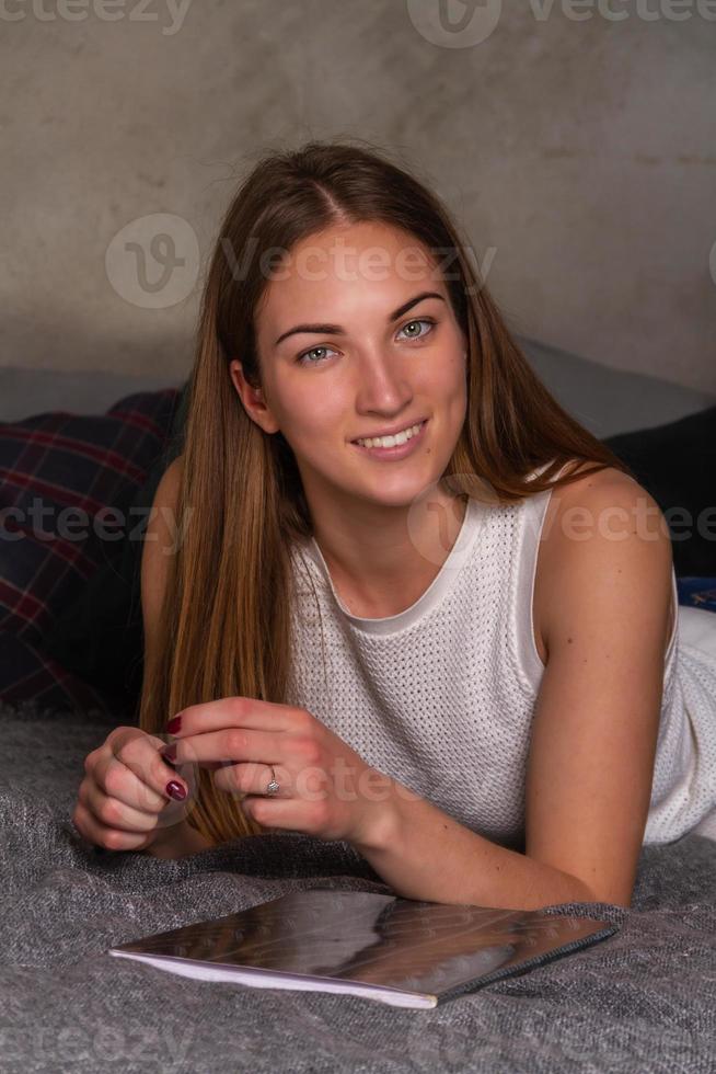 Smiling woman lying on her stomach in bed with a notebook and playing with her hairs photo