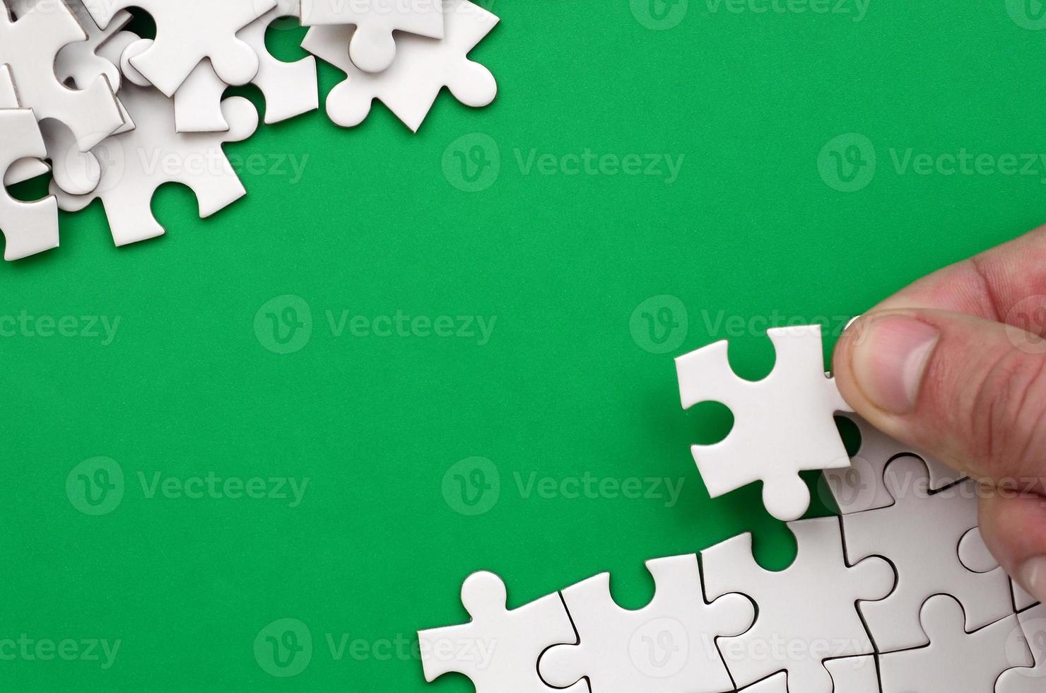 The hand folds a white jigsaw puzzle and a pile of uncombed puzzle pieces lies against the background of the green surface. Texture photo with space for text