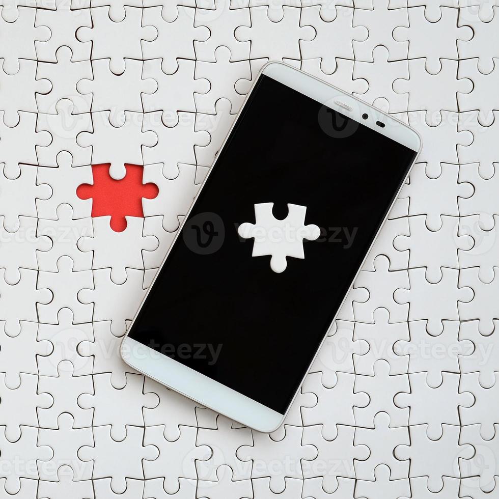 A modern big smartphone with several puzzle elements on the touch screen lies on a white jigsaw puzzle in an assembled state with missing elements photo
