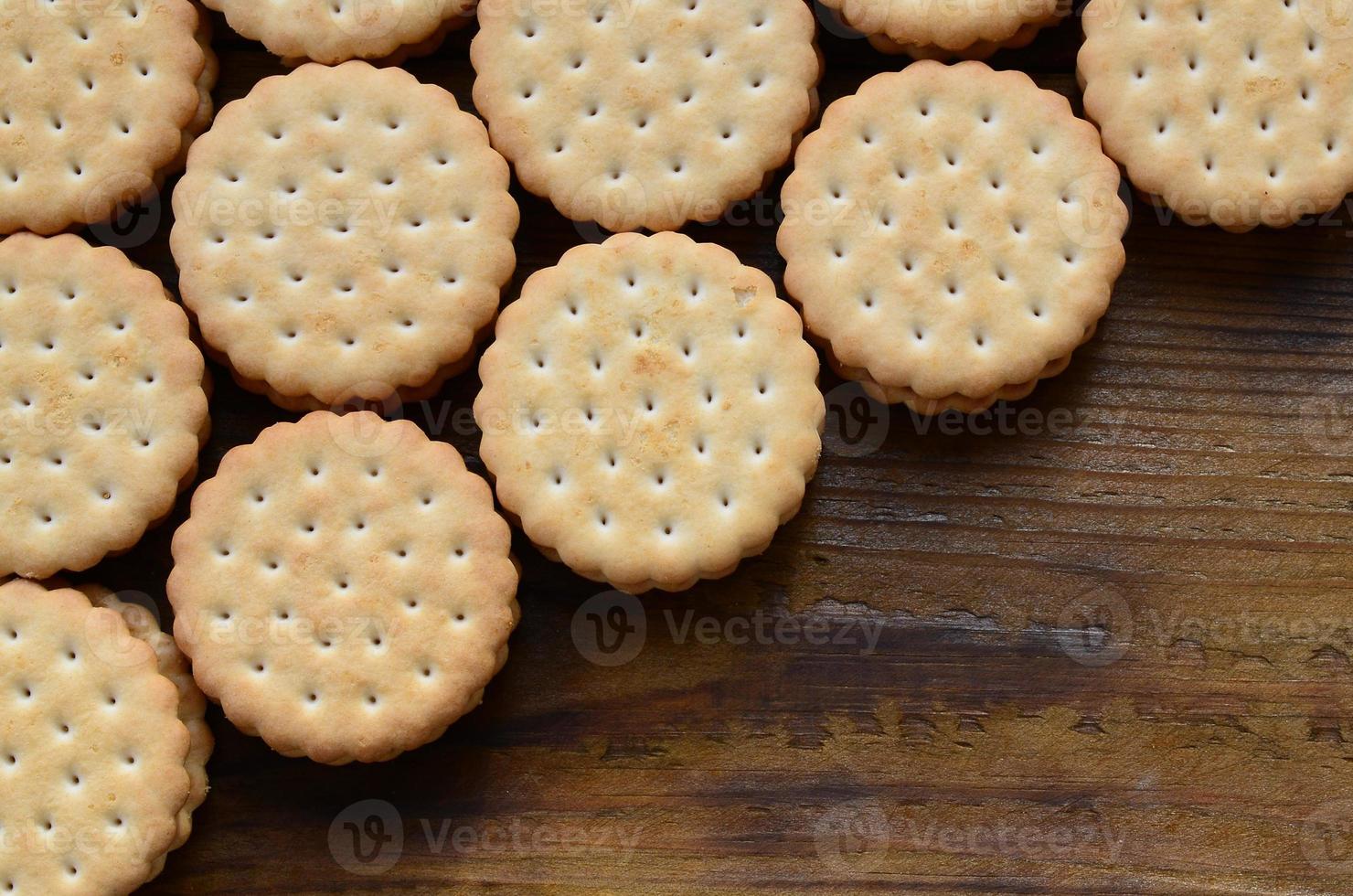 A round sandwich cookie with coconut filling lies in large quantities on a brown wooden surface. Photo of edible treats on a wooden background with copy space