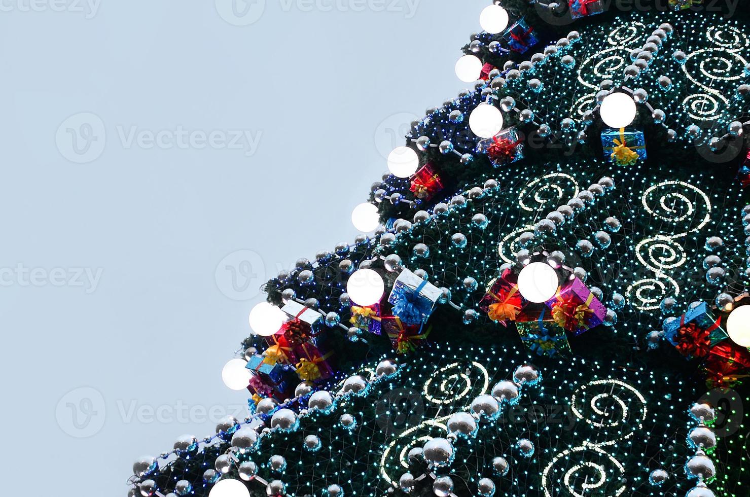 A fragment of a huge Christmas tree with many ornaments, gift boxes and luminous lamps. Photo of a decorated Christmas tree close-up with copy space