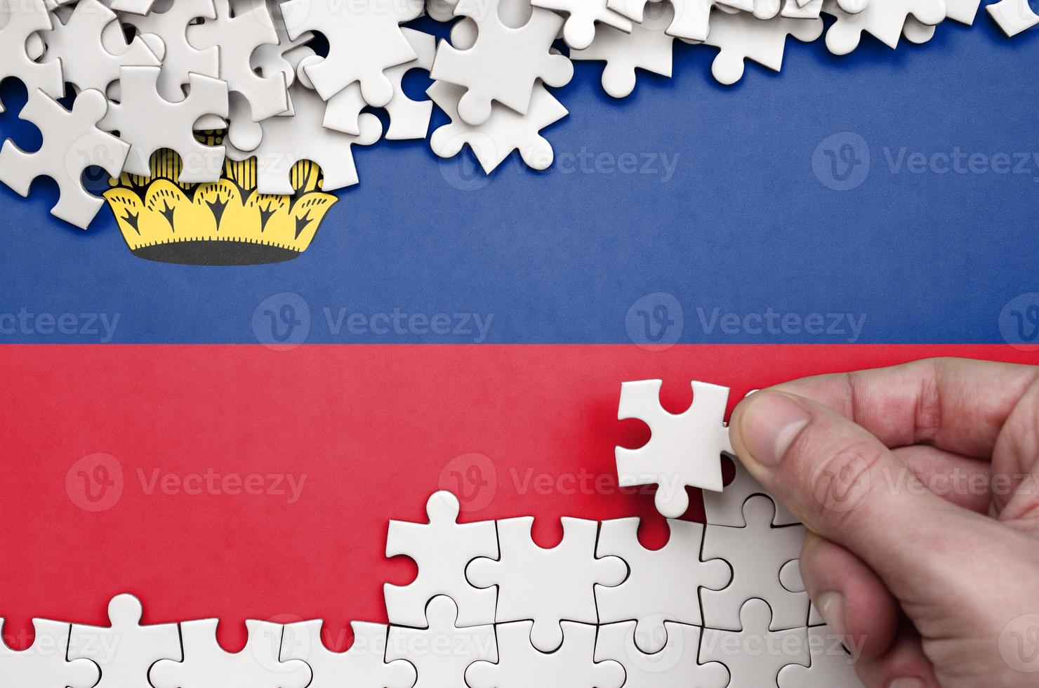 Liechtenstein flag is depicted on a table on which the human hand folds a puzzle of white color photo