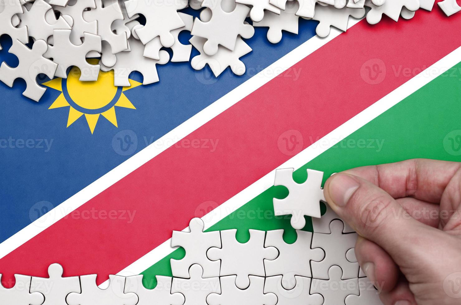 Namibia flag is depicted on a table on which the human hand folds a puzzle of white color photo