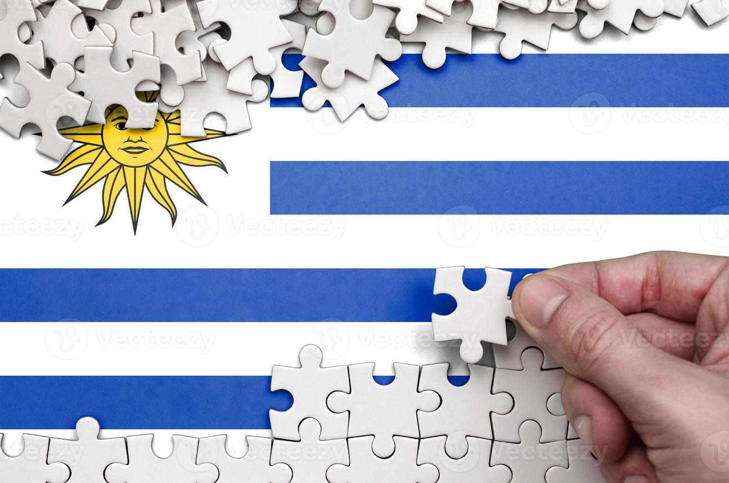 Uruguay flag is depicted on a table on which the human hand folds a puzzle of white color photo