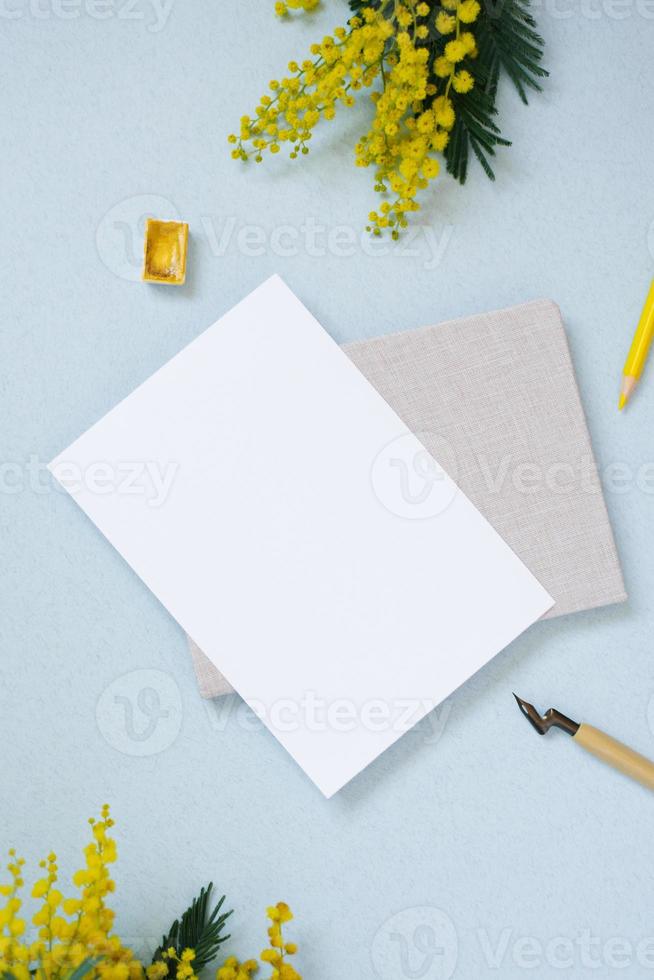 White sheet of paper for text copy space surrounded by spring mimosa, notepad, yellow pencil, watercolors in cuvettes on a blue background. Workplace of a blogger, artist, or calligrapher photo