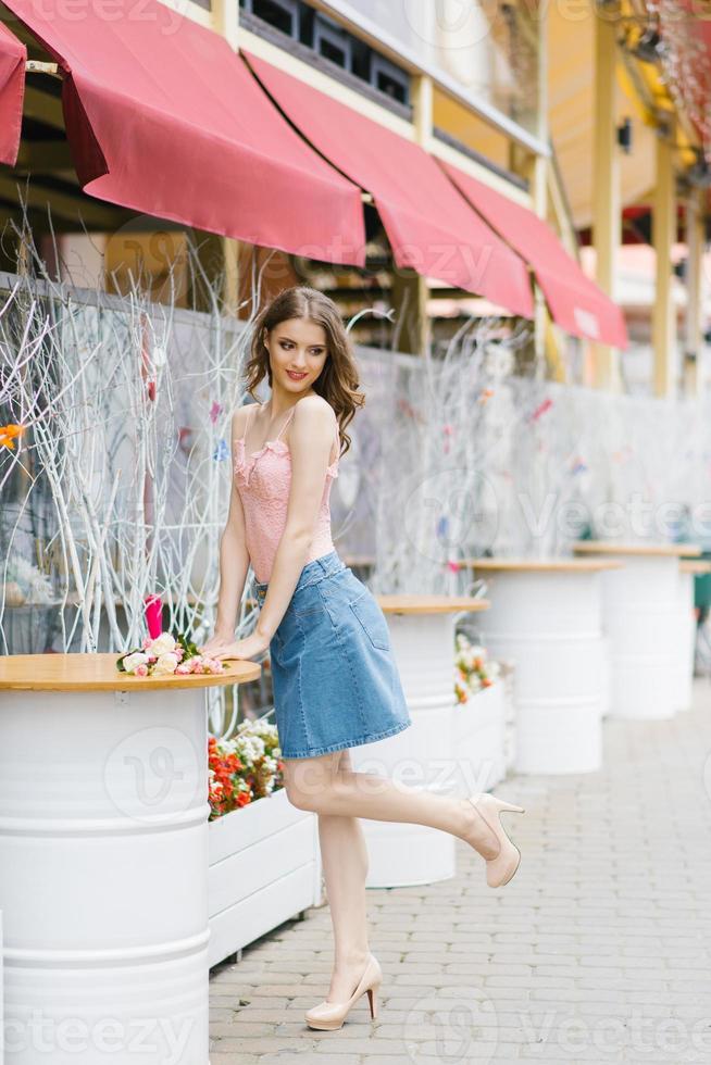 Cute young woman in a denim skirt and pink top stands near a metal white barrel on a walk in the city photo