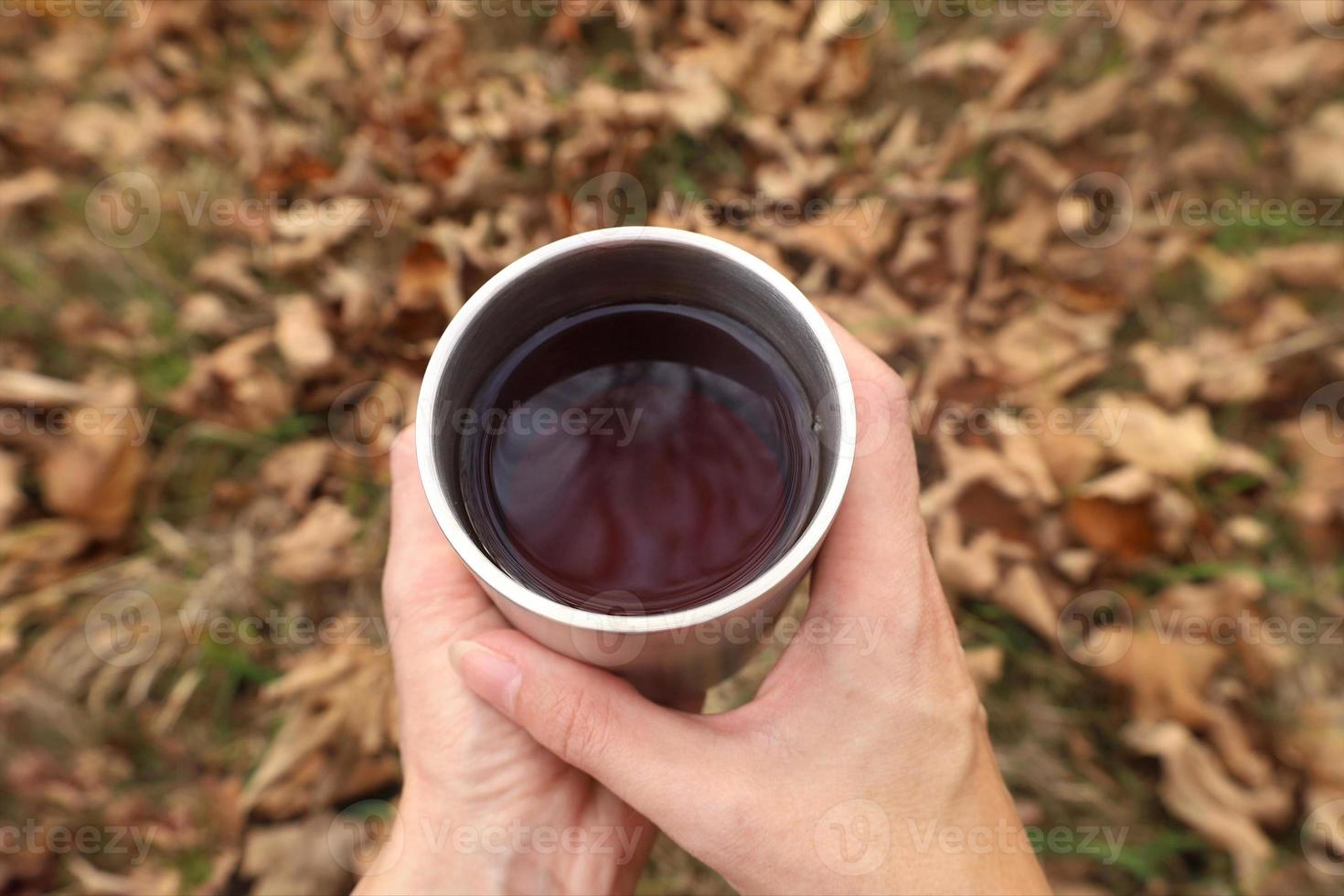 Female hands hold stainless steel cup with hot tea. Selective focus. Blurred fallen foliage on background. Autumn hygge mood. Outdoors, picnic, camping, traveling, trekking concept. photo