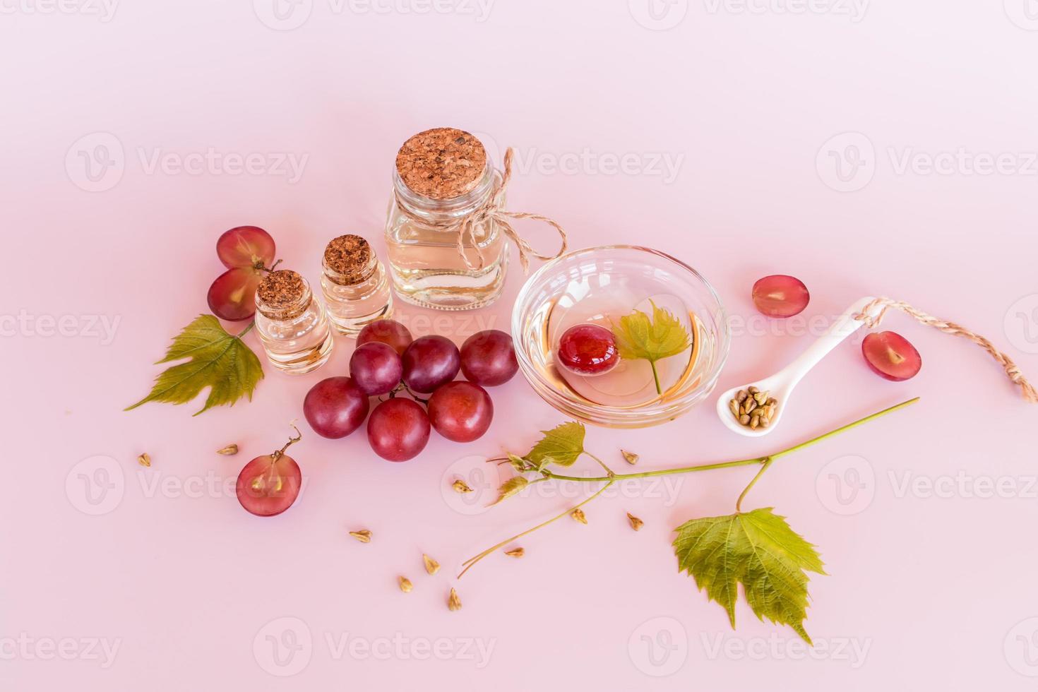 a glass bottle with grape oil, a bowl and a ceramic spoon with grape seeds on a pink background with grape berries. vitamins and minerals. photo