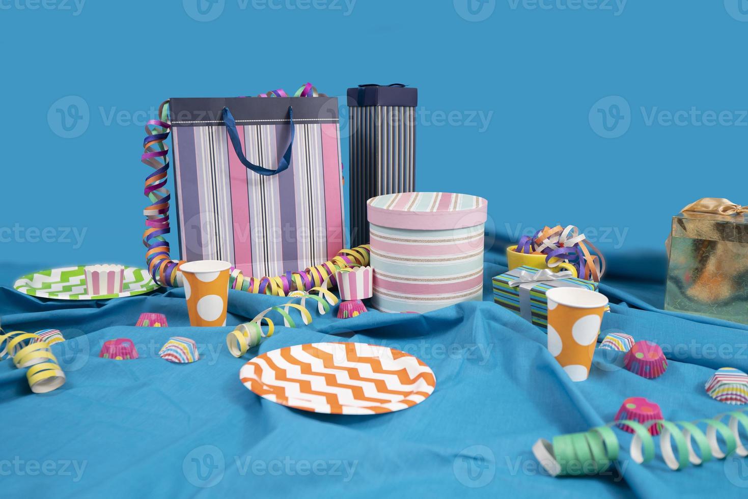 Messy party table with gift bags and boxes, plastic plates and cups on a table with a blue tablecloth against a light blue background photo