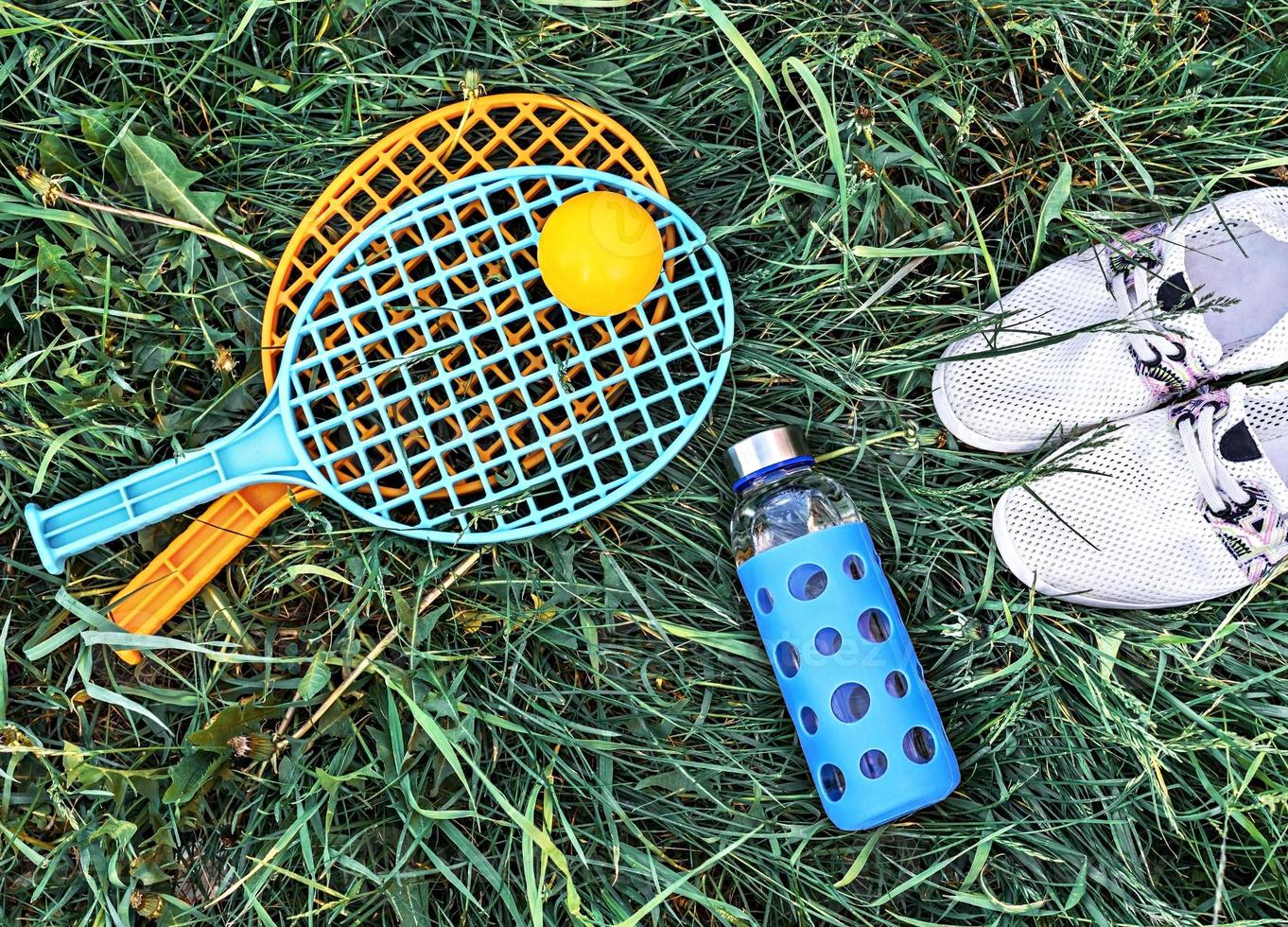 summer flat lay on green grass with rackets and ball, glass bottle of water and summer white shoes photo