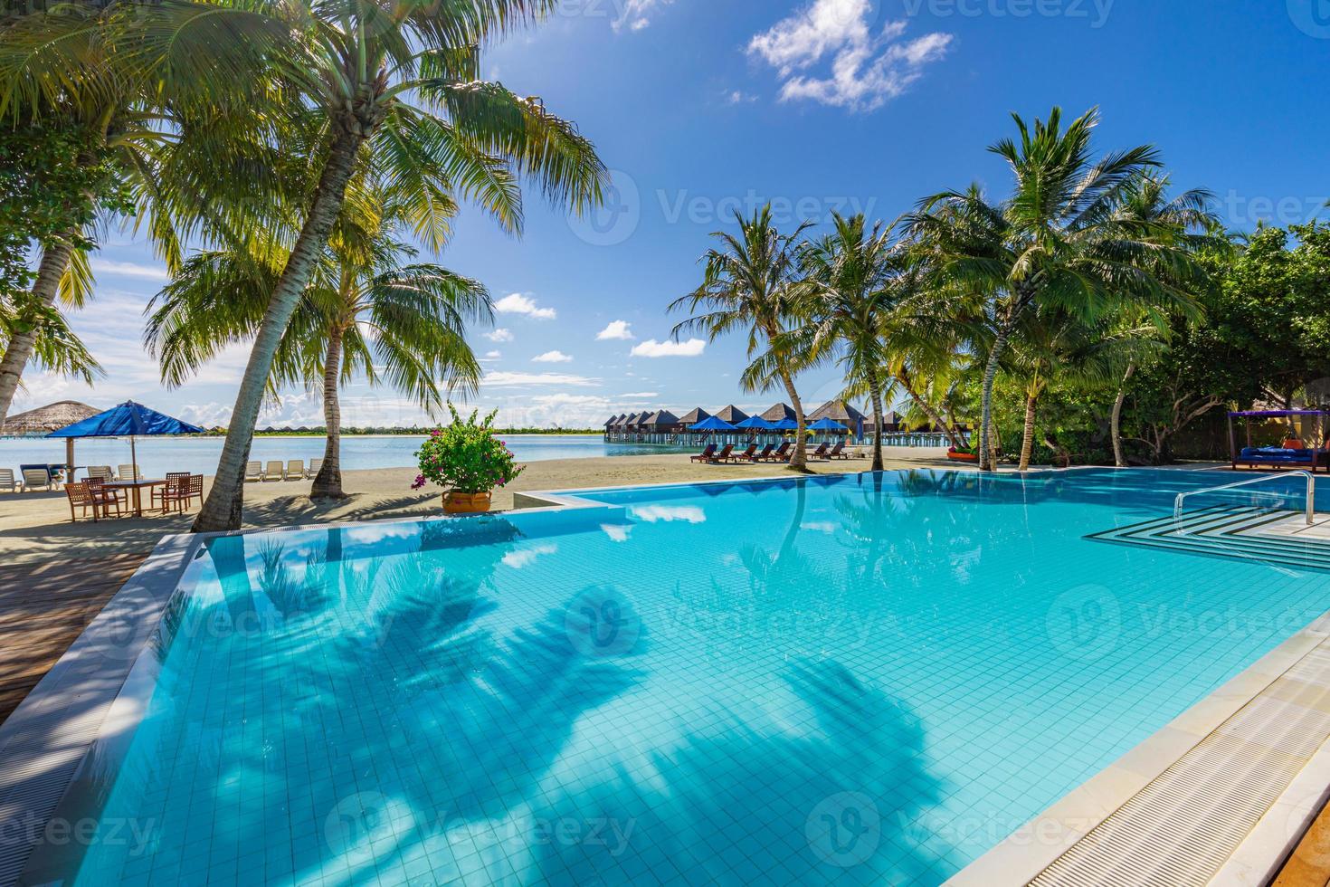 Outdoor tourism landscape. Luxurious beach resort with swimming pool and beach chairs or loungers umbrellas with palm trees and blue sky, sea horizon. Summer island relax travel and idyllic vacation photo