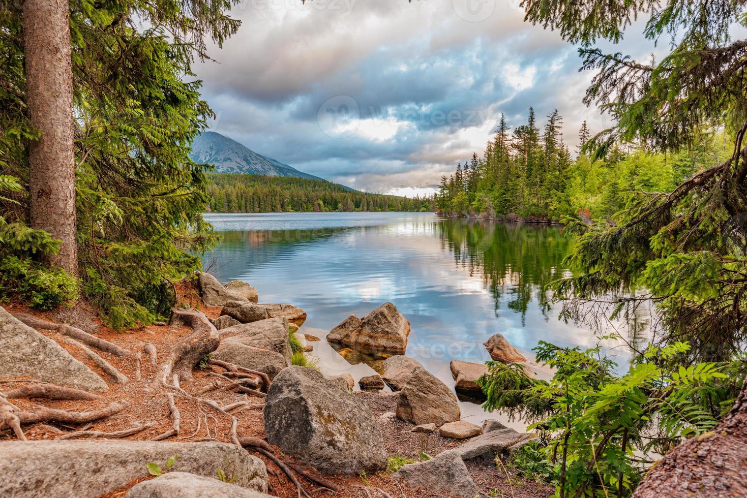 Beautiful mountain landscape, picturesque High Tatra lake in summer morning. Amazing nature panorama, rocks boulders with calm water reflection and pine tree forest. Tranquil scenic view, sunny clouds photo