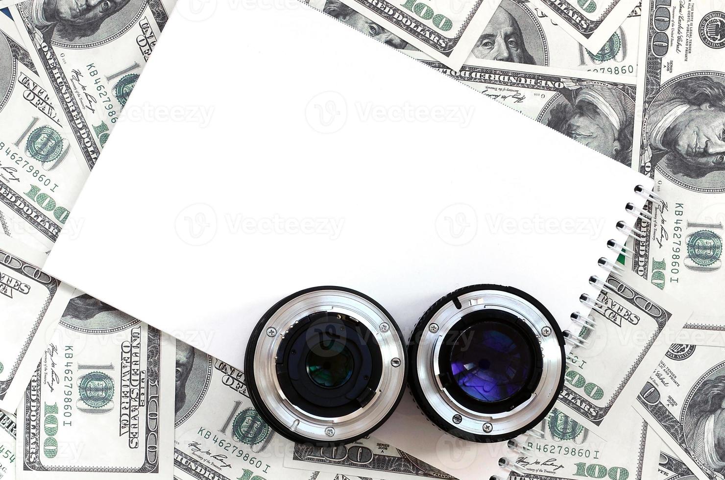 dos lentes fotográficos y un cuaderno blanco se encuentran en el fondo de muchos billetes de dólar. espacio para texto foto