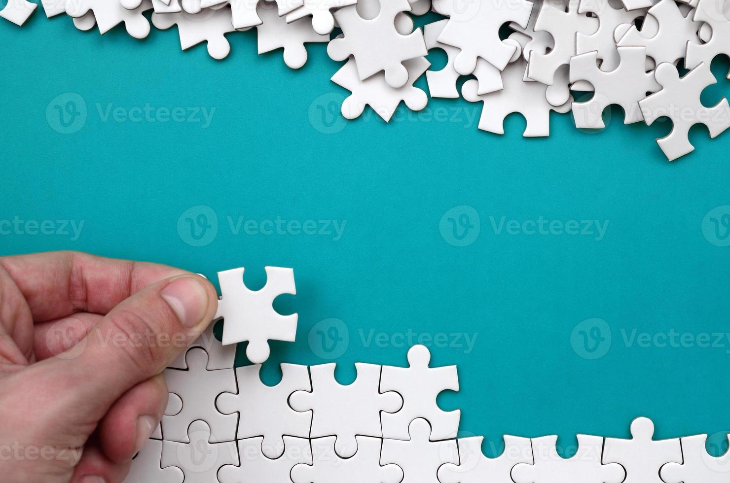 The hand folds a white jigsaw puzzle and a pile of uncombed puzzle pieces lies against the background of the blue surface. Texture photo with space for text