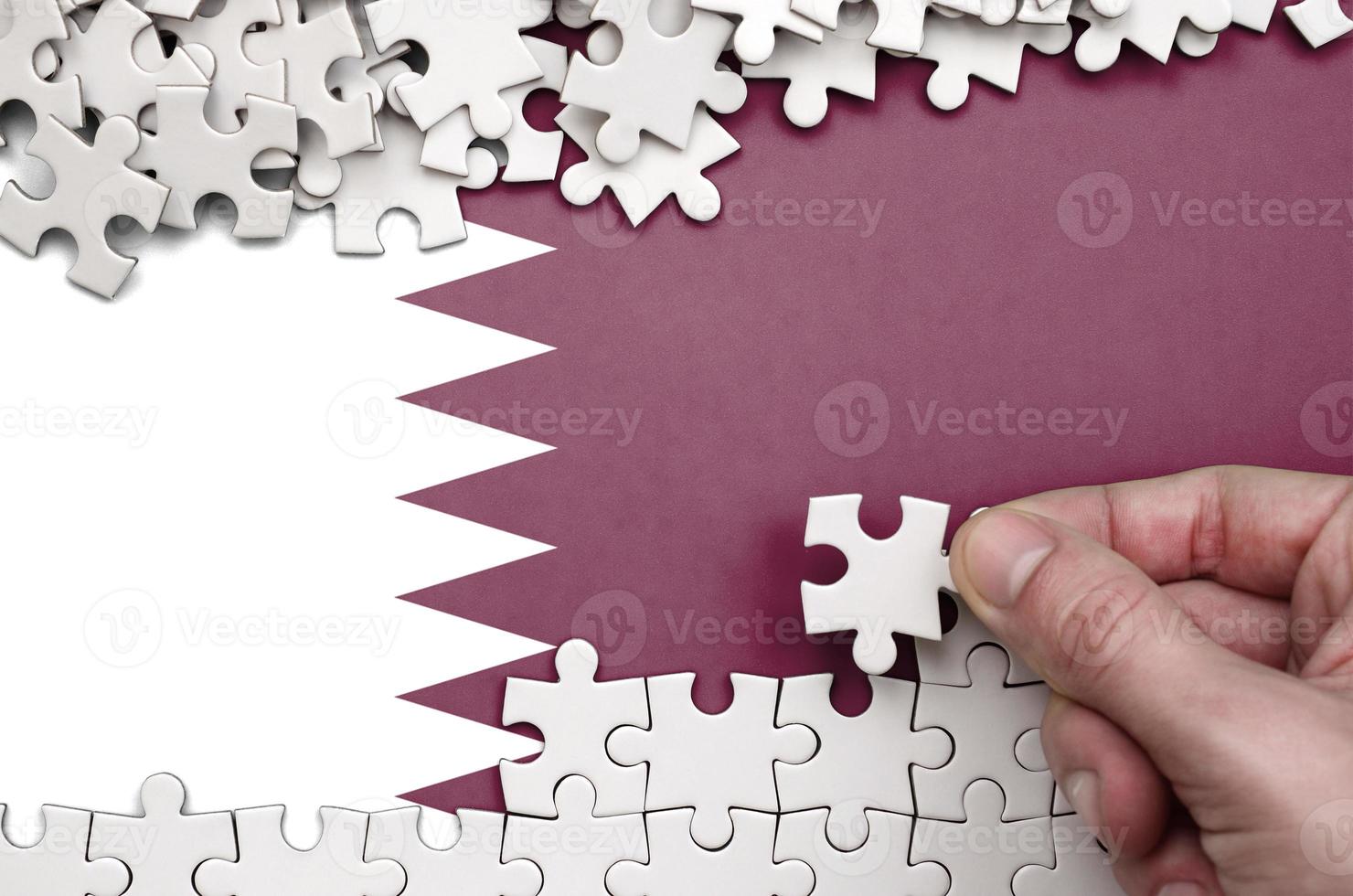Qatar flag is depicted on a table on which the human hand folds a puzzle of white color photo