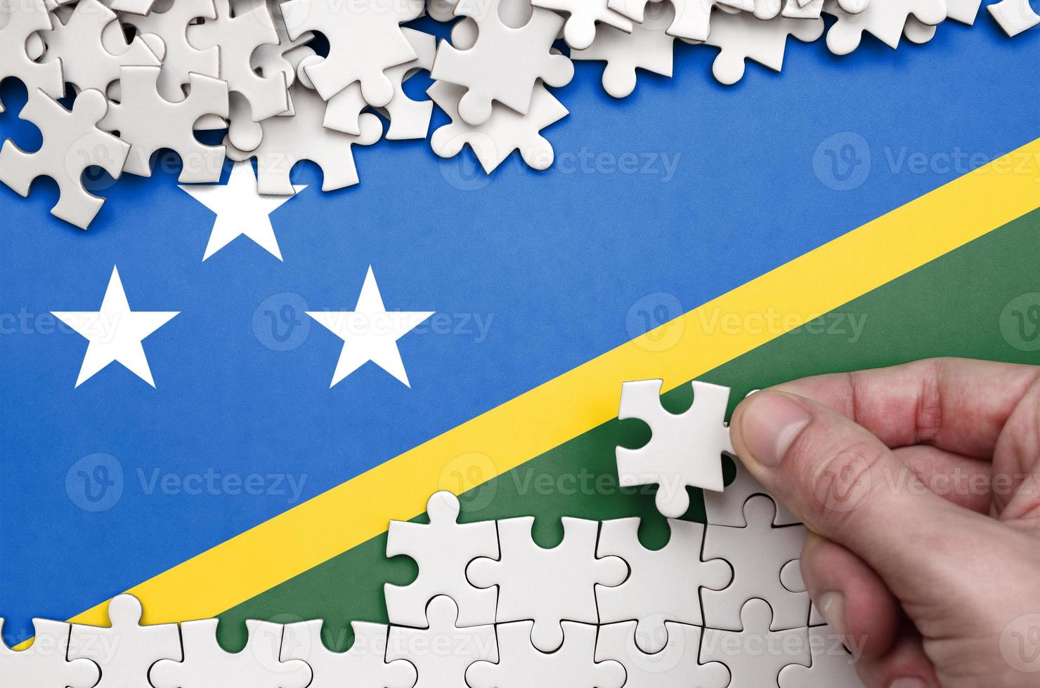 Solomon Islands flag is depicted on a table on which the human hand folds a puzzle of white color photo